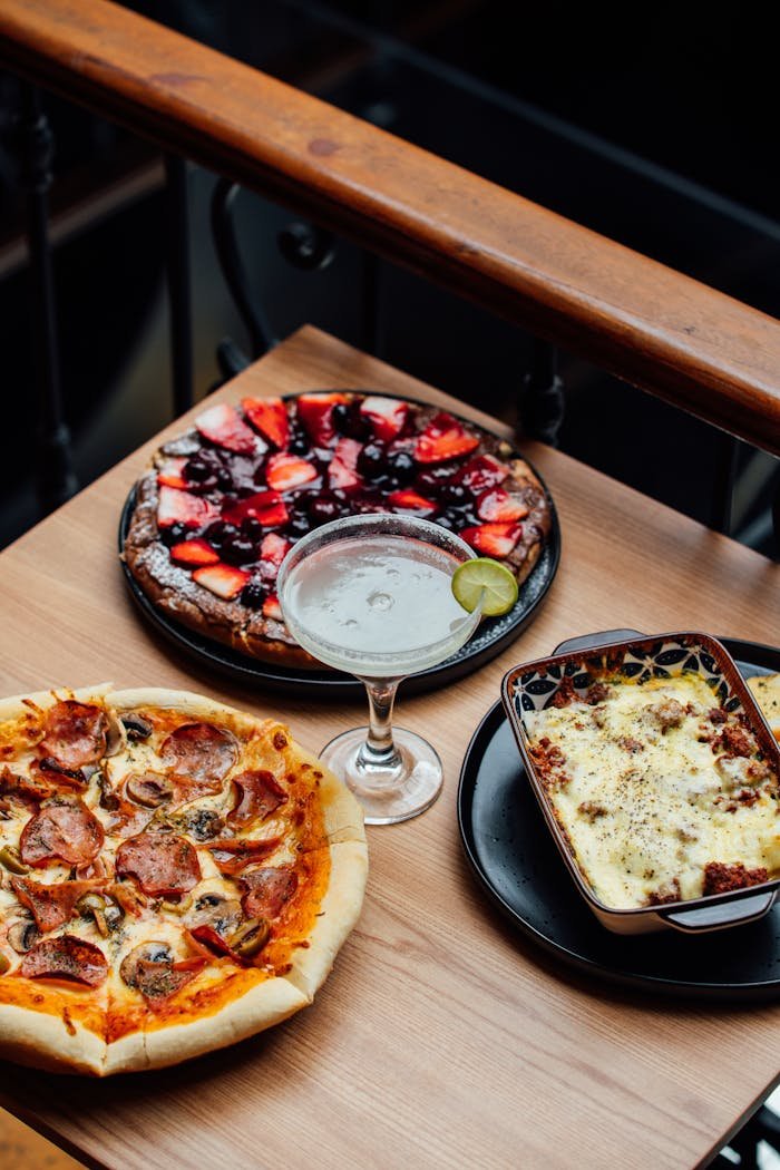 A tempting spread of pizza, dessert pizza, and creamy pasta paired with a cocktail, displayed indoors.