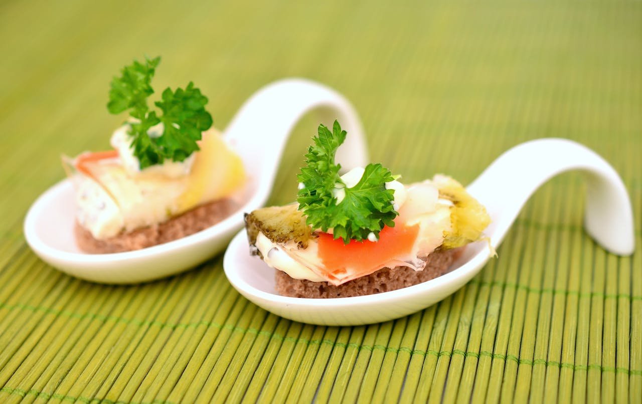 Elegant gourmet appetizers served on ceramic spoons against a bamboo mat background.