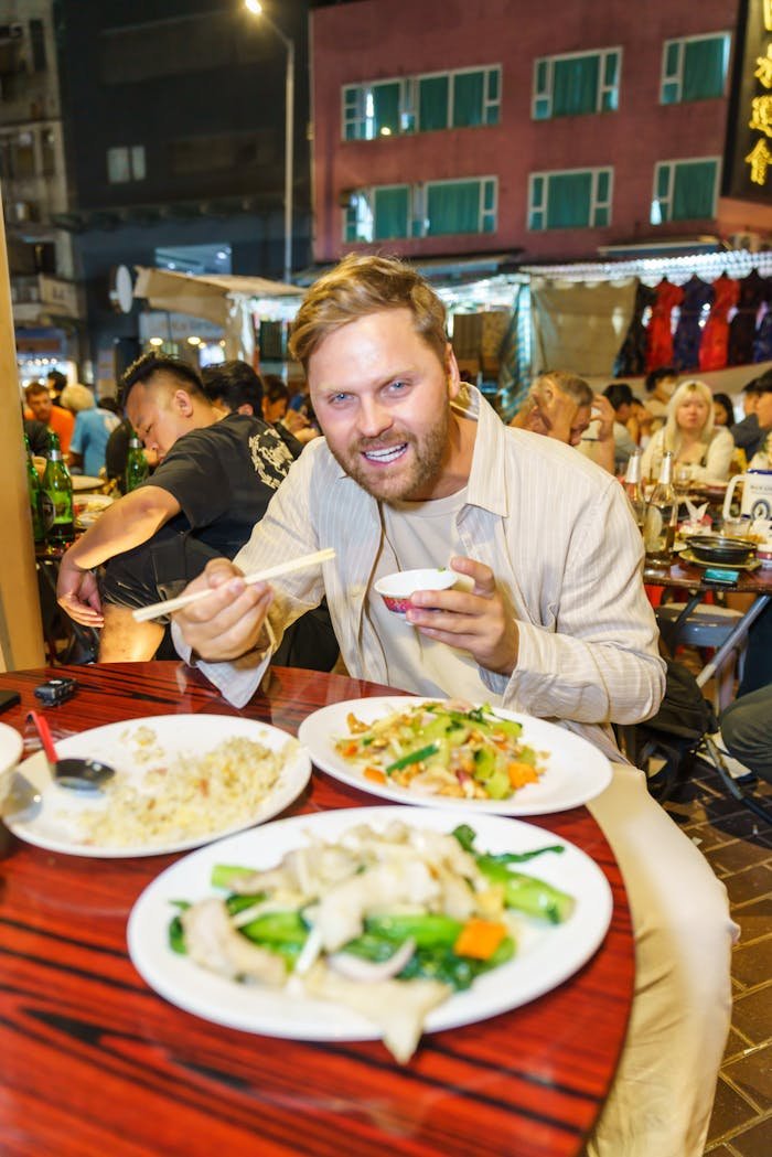 Man dining at an outdoor night market with a vibrant crowd, enjoying Asian cuisine.